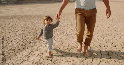 Caucasian baby boy with his father walking barefoot on bottom of dried lake or river, stepping on cracked mud destroyed by global warming - ecological issues concept 4k footage