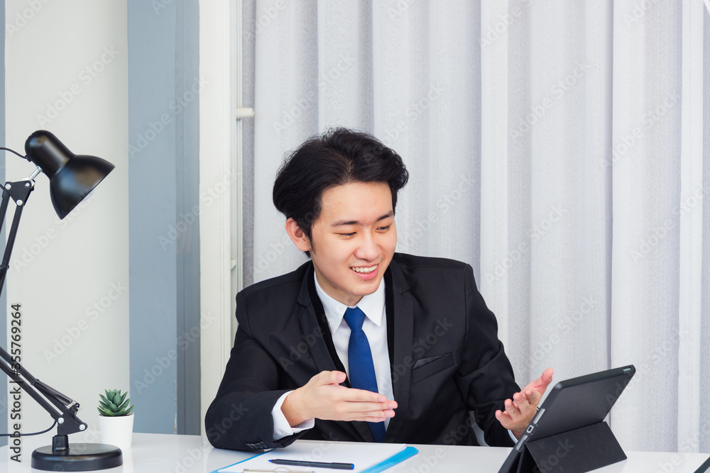 Work from home, Asian young businessman video conference call or facetime he smiling looking to tablet sitting on desk using smart tablet computer raise hand and explain to colleagues at home office Work from home, Asian young businessman video conference call or facetime he smiling looking to tablet sitting on desk using smart tablet computer raise hand and explain to colleagues at home office