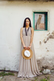 One adult caucasian woman standing in front of the white wall of old traditional house wearing dress looking down sad holding hat alone loneliness solitude and grief concept