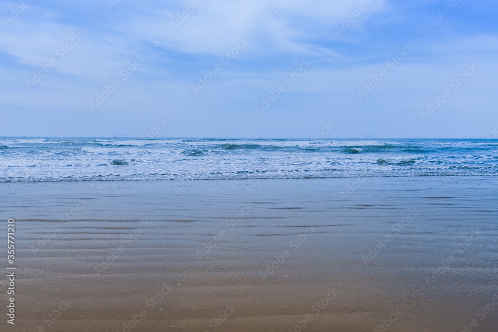 Sandy beach and rough ocean waves and white caps under a blue sky with low wispy cirrus clouds.