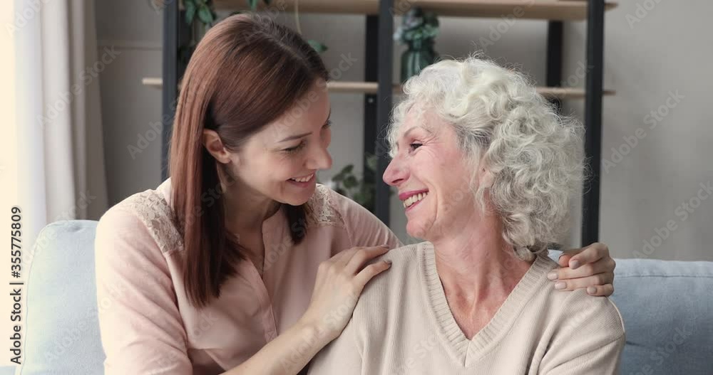 Smiling young beautiful woman touching foreheads with joyful middle aged mother, sitting together on sofa. Affectionate different generations family enjoying sweet tender moment, looking at camera. Smiling young beautiful woman touching foreheads with joyful middle aged mother, sitting together on sofa. Affectionate different generations family enjoying sweet tender moment, looking at camera.