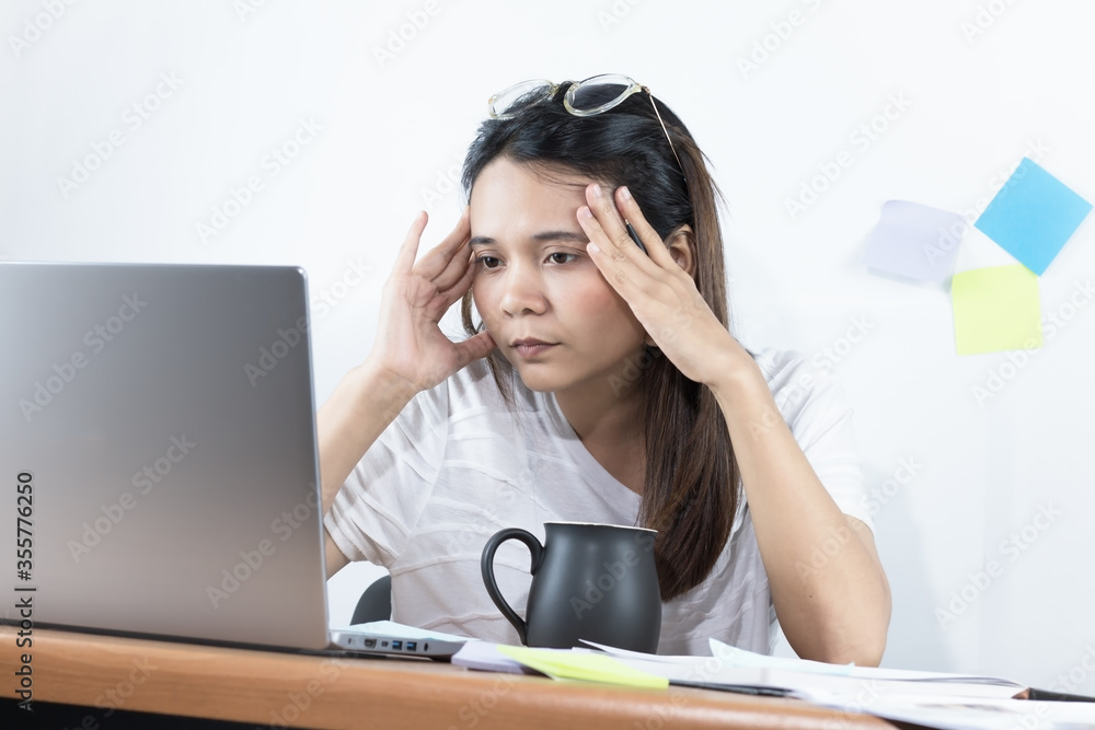 Asian woman wearing a white T-shirt Working at home With serious emotions and thinking power, and hands are holding their heads Eyes looking at the laptop computer screen and the white wall background Asian woman wearing a white T-shirt Working at home With serious emotions and thinking power, and hands are holding their heads Eyes looking at the laptop computer screen and the white wall background