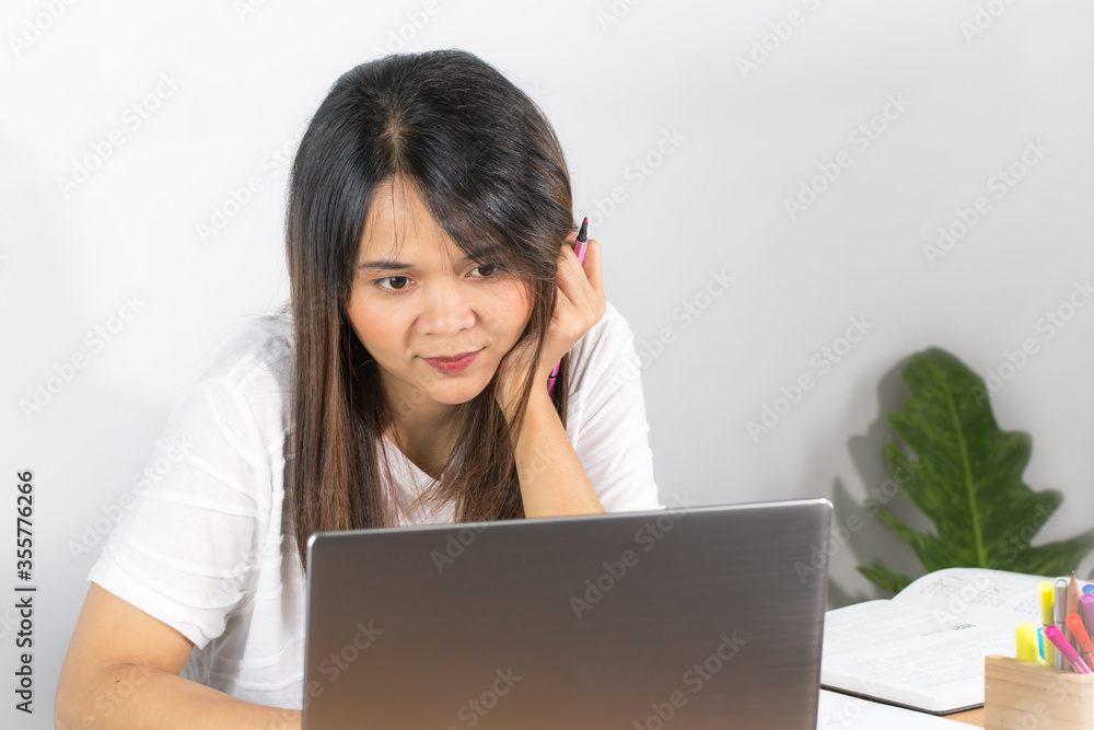 Asian women wear white T-shirt. Sitting happily working at home Looking at the grey laptop screen on the desk and the hand holding the chin and the white wall background Asian women wear white T-shirt. Sitting happily working at home Looking at the grey laptop screen on the desk and the hand holding the chin and the white wall background