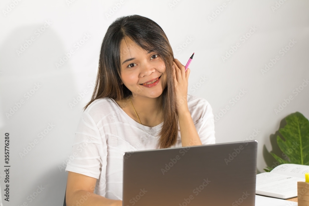 Asian woman wearing white T-shirt Is working happily at home Open the grey notebook screen on the brown wooden office desk and the pen on the desk. Whitewall background and decorated with green trees Asian woman wearing white T-shirt Is working happily at home Open the grey notebook screen on the brown wooden office desk and the pen on the desk. Whitewall background and decorated with green trees