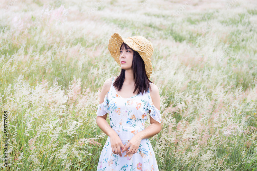 The girl wearing a hat, wearing a white dress, standing in the middle of the grass with beautiful white flowers with a relaxed and happy mood.