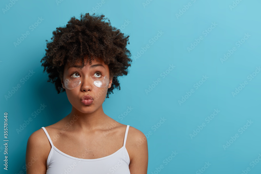 Young beautiful woman with clean fresh facial skin, uses eye patches, keeps lips rounded, enjoys rejuvenation treatment or anti aging therapy, looks above, isolated on blue background, empty space