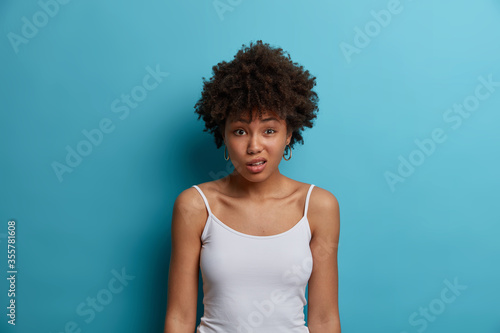 Photos Puzzled displeased woman with Afro hairstyle, frowns face, reacts to negative news, hears unpleasant rumor, wears casual white vest, isolated over blue background