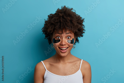 Close up shot of happy dark skinned woman has anti aging eye therapy, applies co Fototapet
