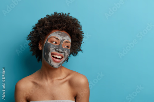 Joyful positive dark skinned curly woman stands naked indoor, applies beauty mud mask for perfect soft facial skin, cares about complexion, looks gladfully away, isolated on blue background.