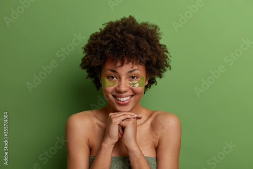 Headshot of positive dark skinned woman enjoys morning skin care routine  applies moisturizing mask patches under eyes  cares about skin  has refreshed happy expression  poses against green background