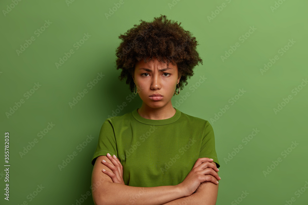 Grumpy offended millennial woman holds hands crossed on chest, frowns face, has quarrel with someone, angry expression, wears green t shirt, being mad at you, feels insulted, unwilling to talk Grumpy offended millennial woman holds hands crossed on chest, frowns face, has quarrel with someone, angry expression, wears green t shirt, being mad at you, feels insulted, unwilling to talk