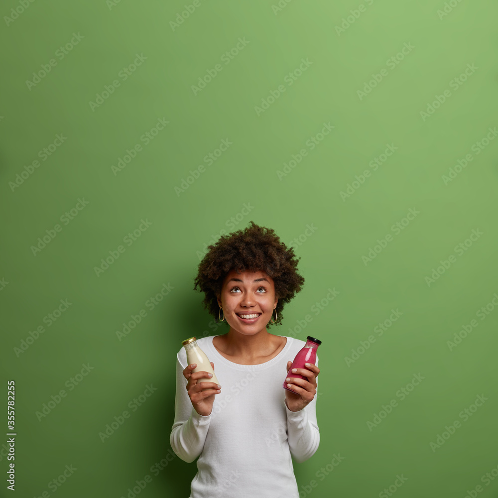 Positive curly woman holds smoothie and vegan milk, drinks detox beverage, has balanced proper nutrition, concentrated above with broad smile, sees something pleasant, isolated on green background Positive curly woman holds smoothie and vegan milk, drinks detox beverage, has balanced proper nutrition, concentrated above with broad smile, sees something pleasant, isolated on green background