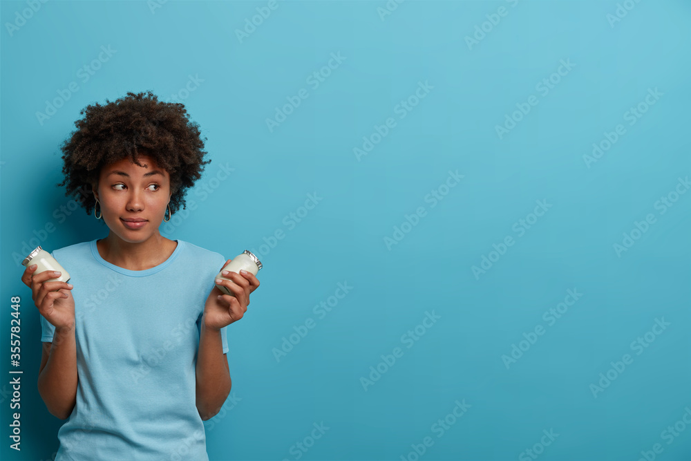 Healthy sporty woman with Afro hair holds glass jars of fresh yoghurt, going to prepare breakfast, has proper nutrition, concentrated aside, wears casual clothes, poses against blue background