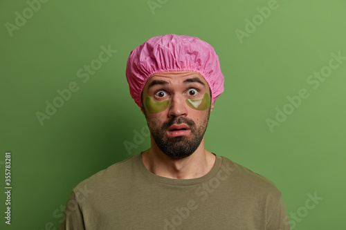 Young stunned unshaven European man stares with disbelief, finds out shocking news, wears bath cap and green undereye patches for reducing wrinkles and puffiness, isolated on green background