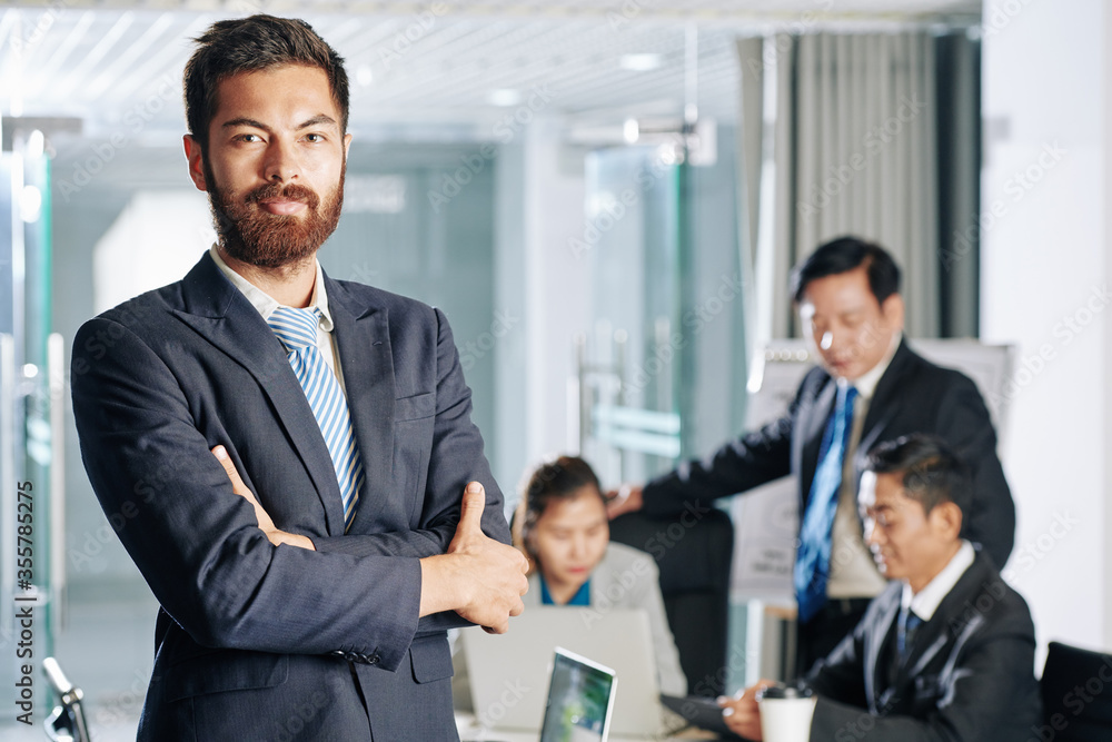 Portrait of confident positive young entrepreneur, his colleagues development anti crisis measures in background Portrait of confident positive young entrepreneur, his colleagues development anti crisis measures in background