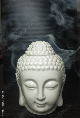 A close up shot of a ceramic sculpture based on buddha face with eyes closed, placed on a dark brown wooden table with light smoke effects in the background.