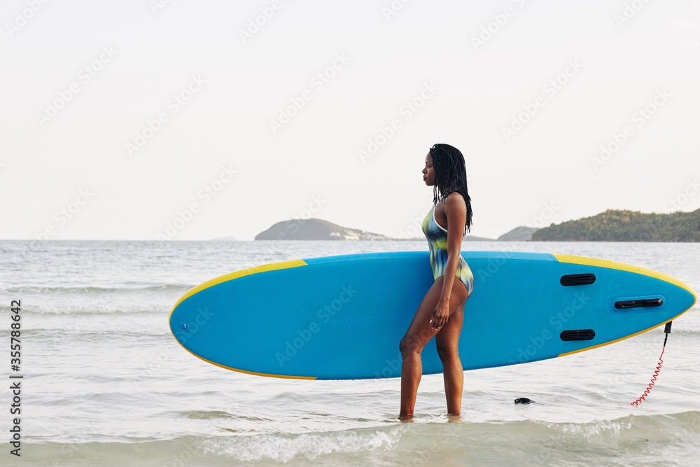 Serious pretty fit young woman carrying sup board and looking at sea waves