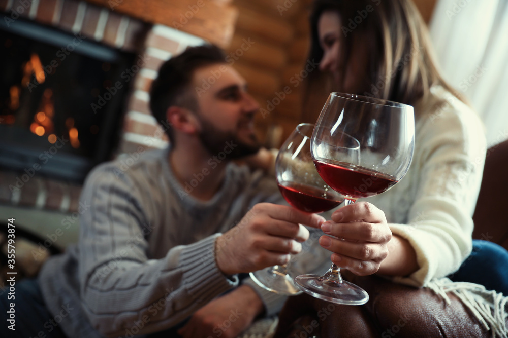 Couple with glasses of wine near fireplace indoors, focus on hands. Winter vacation