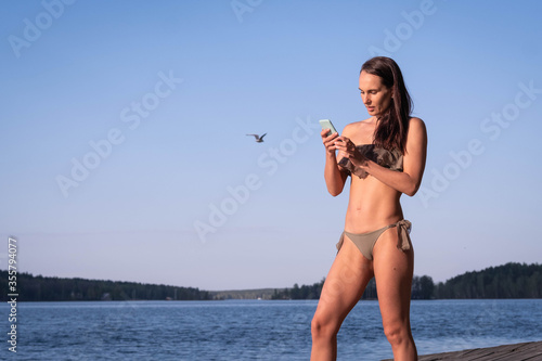 A smiling beautiful woman writes a message on her smartphone and stands on the pier, with a seagull flying behind her