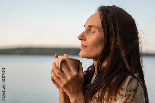 Portrait of a beautiful girl with dark long hair inhaling the aroma of coffee from a ceramic mug in the early morning