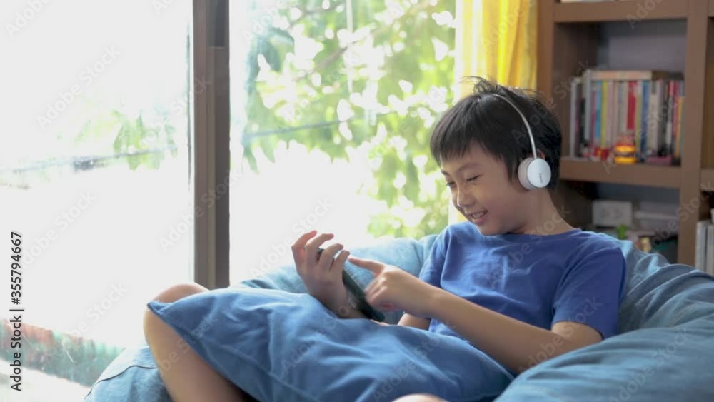 Happy young asian boy while connection on blue headphones is using a smartphone and smiling happy while listening to music on the bed at home Happy young asian boy while connection on blue headphones is using a smartphone and smiling happy while listening to music on the bed at home