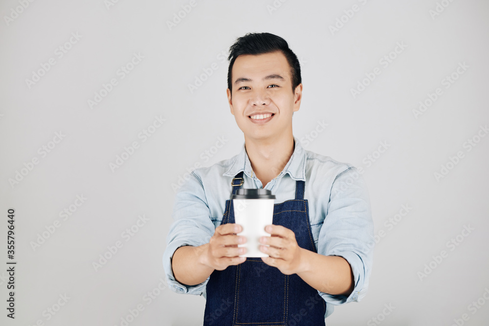 Cheerful young Asian barista giving cup of take away coffee, isolated on grey Cheerful young Asian barista giving cup of take away coffee, isolated on grey