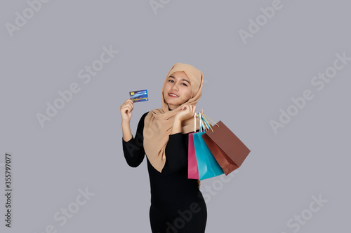 Young asian muslim woman smiling wearing hijab head scarf hand holding credit card and shopping bags gray background.