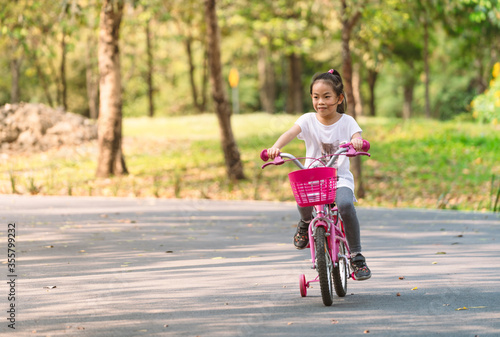 Front view Asian child girl practicing biking bicycle with one supporter wheels. Happy with bicycle, smiling face girl, 6 years old girl. Blurred background tree in park, space for text and design.