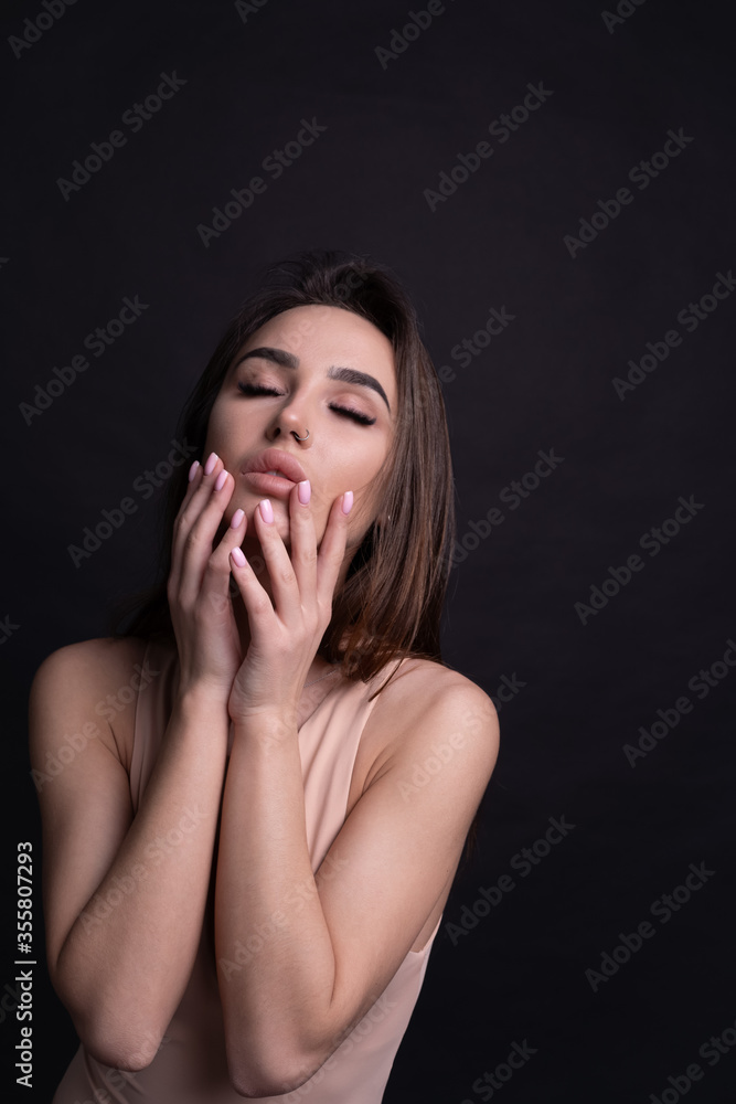 Studio portrait of a young attractive girl closing her eyes and touching her face with both hands emotionally. Nude makeup, beige bodysuit, black background. Studio portrait of a young attractive girl closing her eyes and touching her face with both hands emotionally. Nude makeup, beige bodysuit, black background.