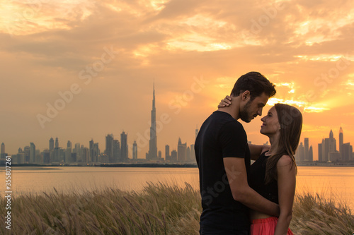 Canvas Print Two lovers on holiday embracing and about to kiss during a beautiful orange sunset sky against a the city view background