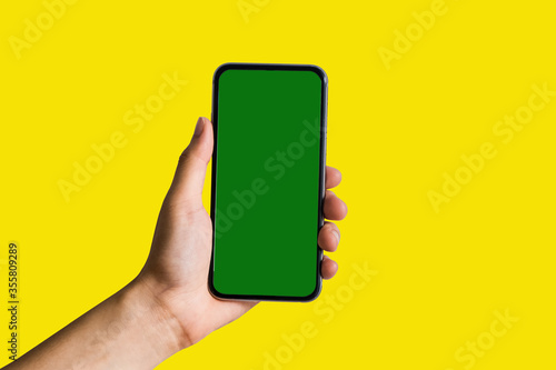 Hand holding white mobile phone with blank white screen in yellow background.