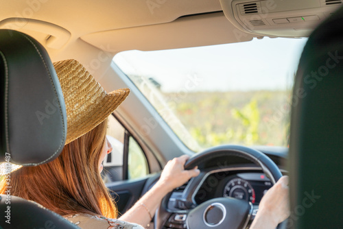 A woman in a hat and a colored dress is sitting at the wheel of a car, the view from behind, the window shows the field.