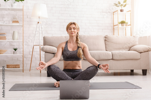 Yoga and meditation at home. Woman in lotus pose, sitting on mat in front of laptop