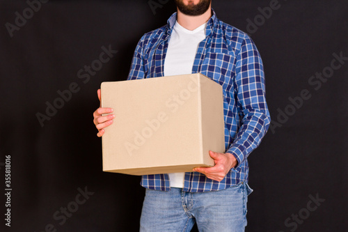 Young parcel delivery man holding a cardboard box in his hands on a black background © fototvv