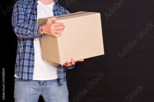 Young parcel delivery man holding a cardboard box in his hands on a black background