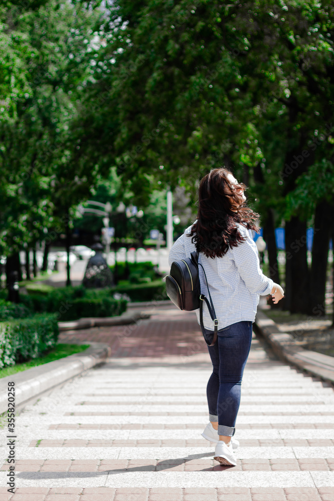Young woman in blue jeans, blue and white square shirt with black backpack, walking in city park. Back shot of brunette girl wondering on the street. Image of young woman. Outdoor activity, lifestyle