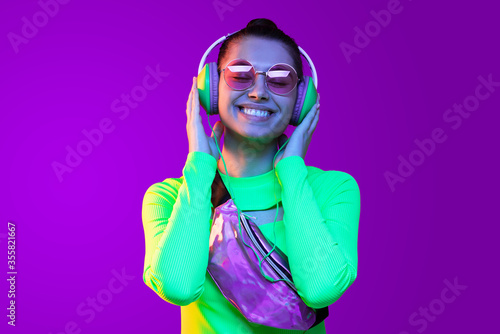 Close-up portrait of young happy girl in neon green top, wearing belt bag, glasses and headphones, listening to favorite song with smile, isolated on purple background