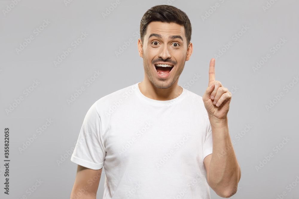 Young handsome guy in white t-shirt feeling deeply amazed and excited by commercial offer, pointing to it up with finger, isolated on gray background Young handsome guy in white t-shirt feeling deeply amazed and excited by commercial offer, pointing to it up with finger, isolated on gray background