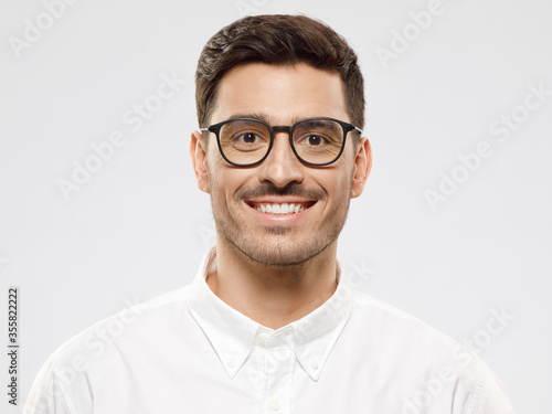 Young handsome smiling man wearing glasses and white shirt, isolated on gray background. Eyewear fashion or optics ads © Damir Khabirov