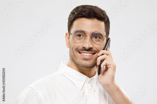 Portrait of young handsome business man in white shirt and trendy round glasses talking on phone, smiling and looking away, isolated on gray background