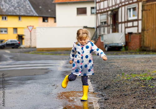 Little toddler girl wearing yellow rain boots, running and walking during sleet on rainy cloudy day. Cute happy child in colorful clothes jumping into puddle, splashing with water, outdoor activity