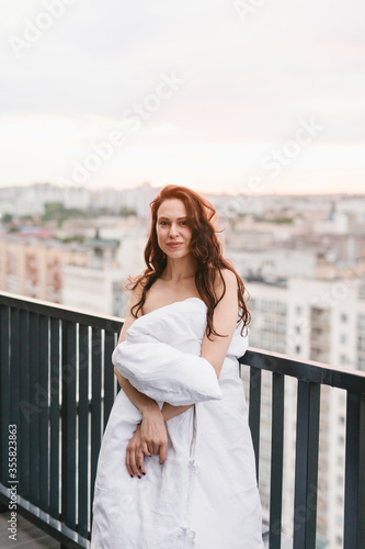 Beautiful girl on the terrace in a white blanket at sunset. Girl posing background fashion. Mood girl wrapping with blanket on balcony. Terrace mood.