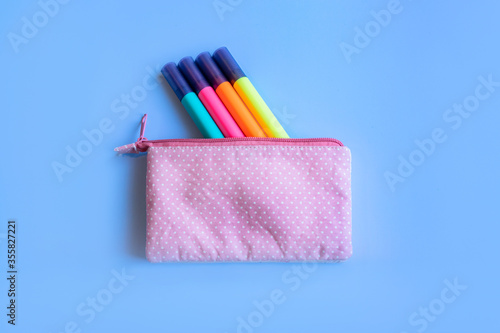 Four markers to highlight text peek out from a pink to speckled pencil case on a blue background. School accessories.