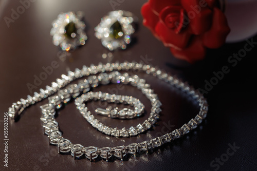 silver chain with precious stones on a brown table, against a background of red rose and earrings with stones