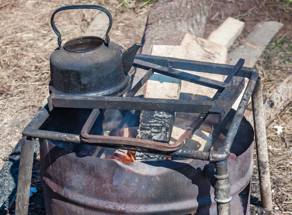 Sooty kettle on a metal stand on the fire from the firewood in a barrel against the ground in summer
