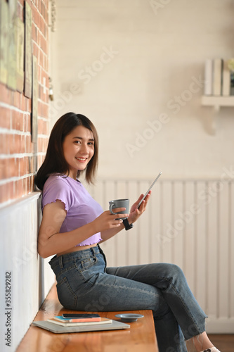 A beautiful woman is holding a coffee cup in her hands while using a computer tablet.