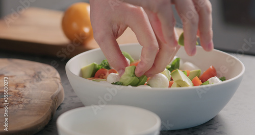 man add avocado into salad with kale, mozzarella and tomatoes in white bowl on kitchen countertop