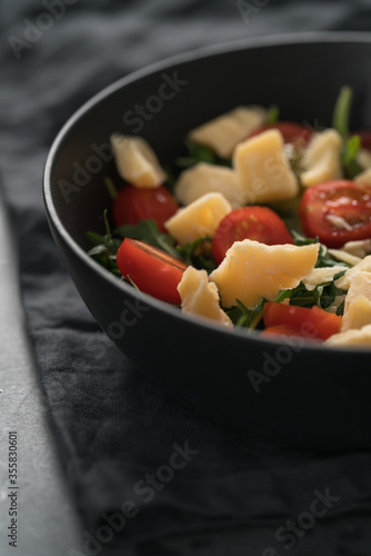 Fresh salad with cherry tomatoes, arugula and hard vintage cheese in black bowl on linen napkin