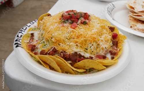 Vászonkép Layers of nachos on a Styrofoam plate piled with hotdogs, salsa, jalapeno and topped with shredded cheese and sliced tomatoes