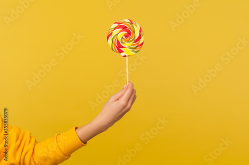 Closeup of hand holding appetizing round rainbow candy on stick, big lollipop in arm, confectionery advertising, glucose sweet foods, sugary dessert. indoor studio shot isolated on yellow background